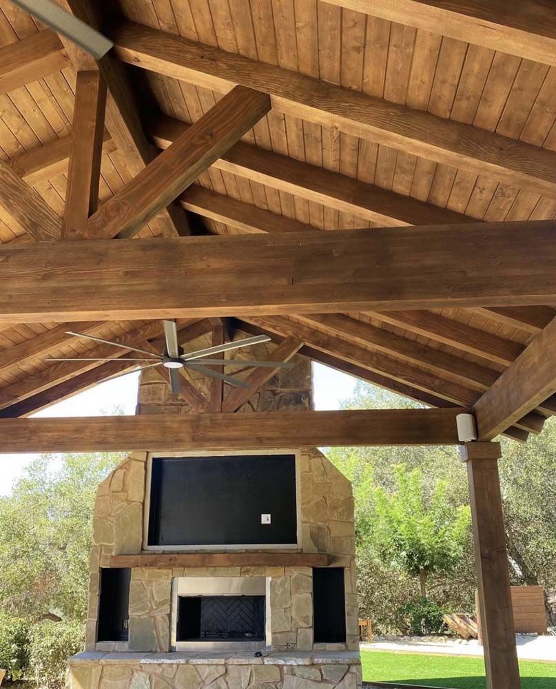 Outdoor wooden pavilion with stone fireplace, TV, and ceiling fan. Brown wood beams and natural stone.