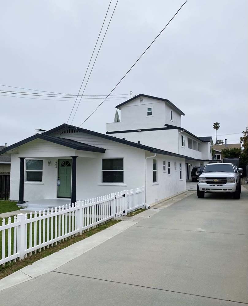 White house with a white picket fence, a long driveway, and a parked SUV on a cloudy day.