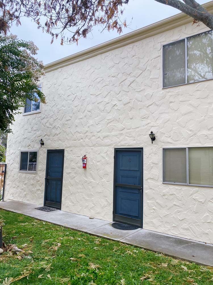Two-story stucco apartment building with blue doors and windows, fire extinguisher, and green grass.