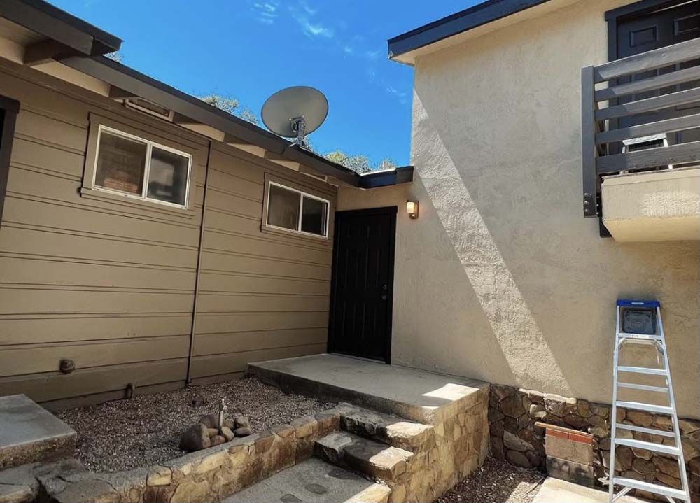 Exterior view of a building with a black door, windows, and a satellite dish. A ladder leans against the wall.