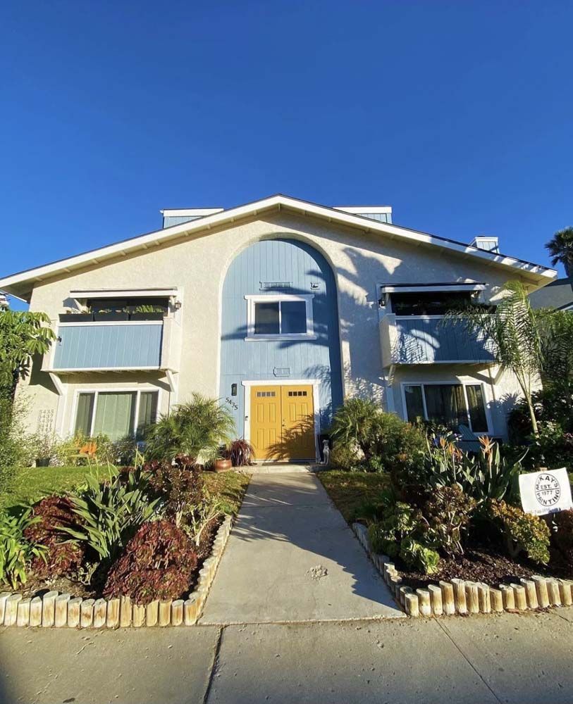 Two-story house with blue and beige facade, yellow door, awnings, and landscaped yard under a clear blue sky.