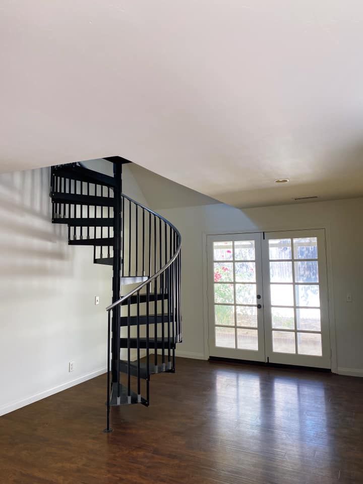 Black spiral staircase in a room with light brown flooring, white walls, and French doors with a colorful pane.