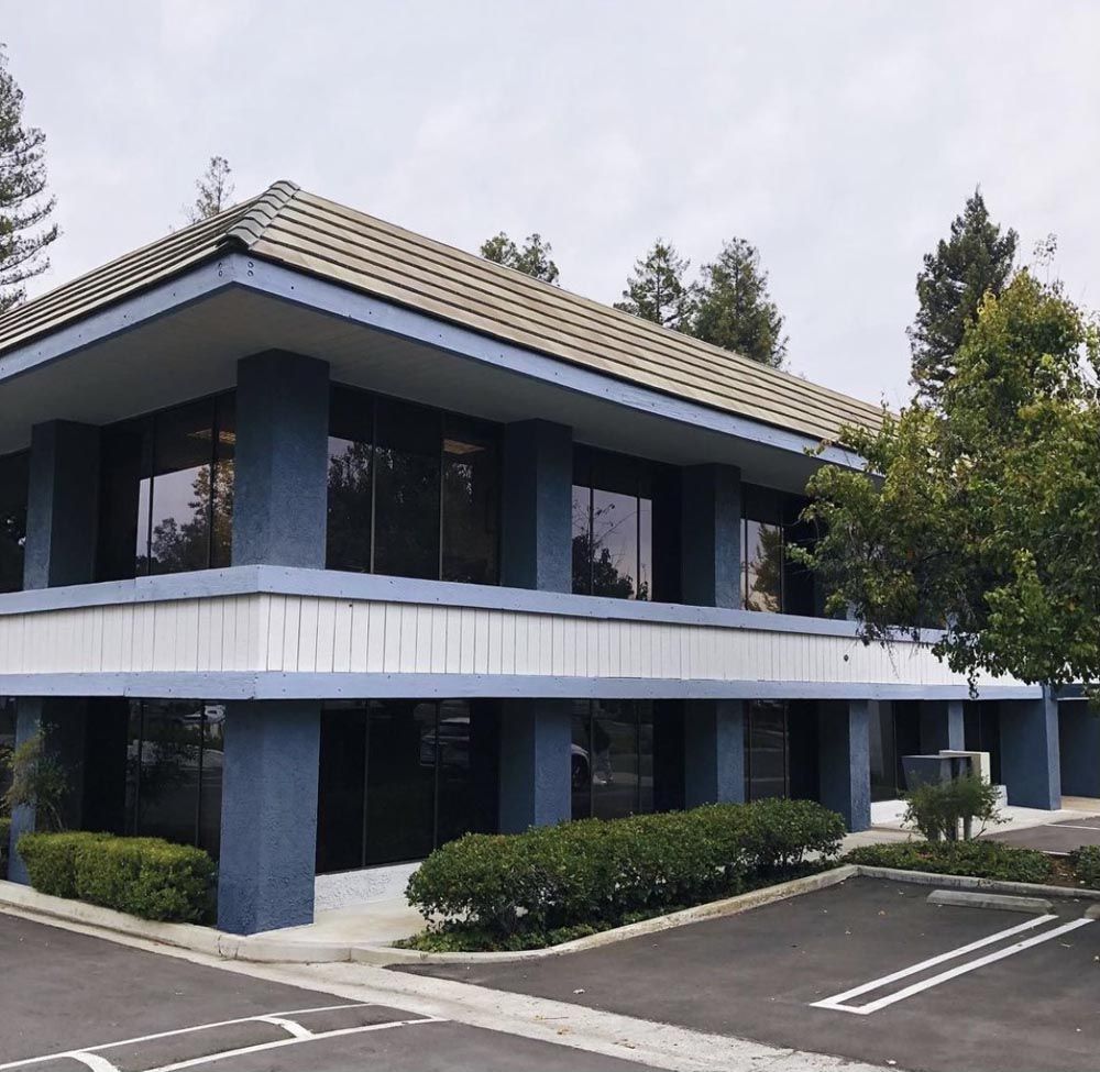 Two-story blue and white office building with dark windows and a tile roof, trees in the background.