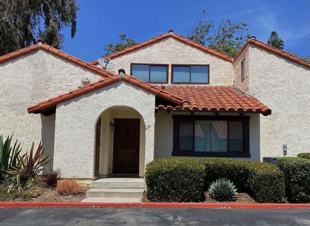 Tan stucco townhouse with red tile roof, dark-framed windows, and neatly trimmed bushes under a clear blue sky.