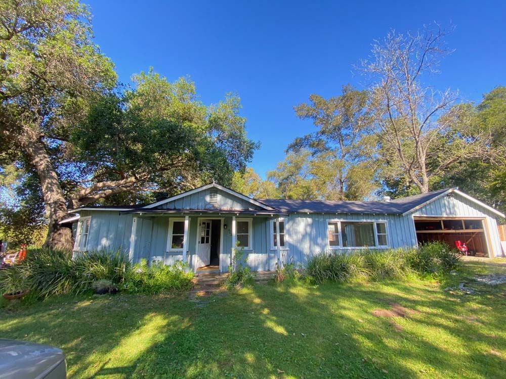 Blue house with open garage door under a blue sky, surrounded by green trees and grass.