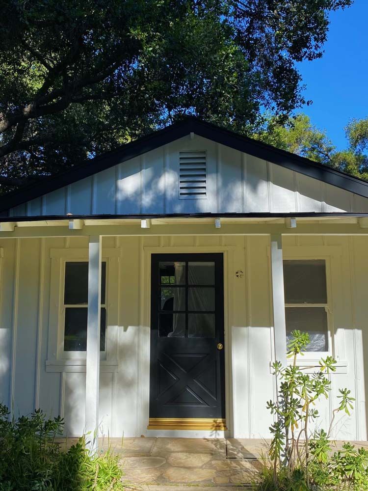 White cottage with black door and roof, porch with two pillars, and lush greenery.