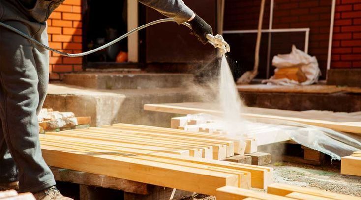Person spraying preservative on lumber outdoors.