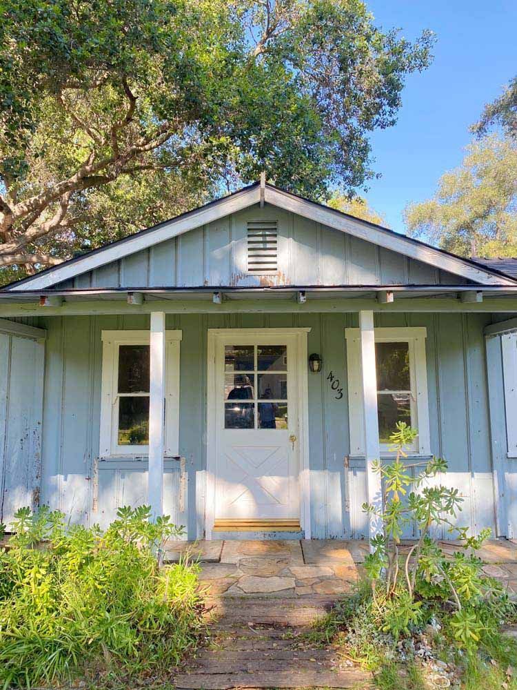 Light blue cottage with white trim, door, and windows, flanked by two posts. Pathway to the entrance.