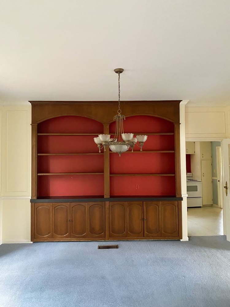 Built-in wooden bookcase with red backing and a chandelier, situated in a room with light blue carpet.