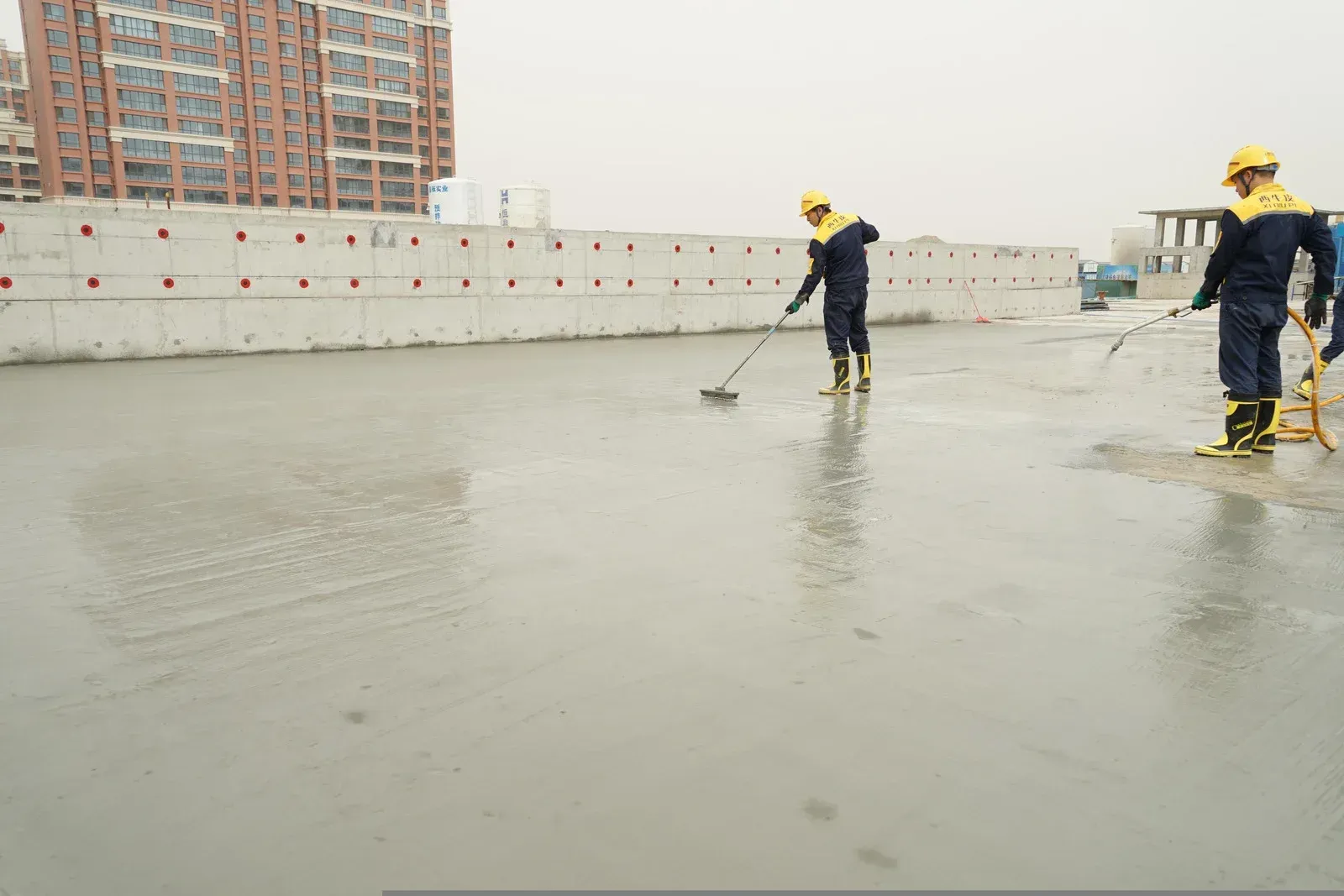 Two construction workers in yellow helmets smoothing wet concrete with tools. Outdoor setting.
