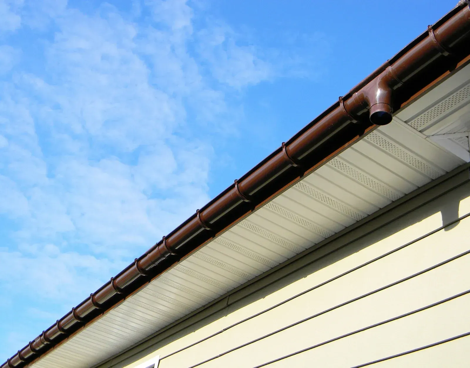 Brown gutters on a cream-colored house against a blue sky with light clouds.