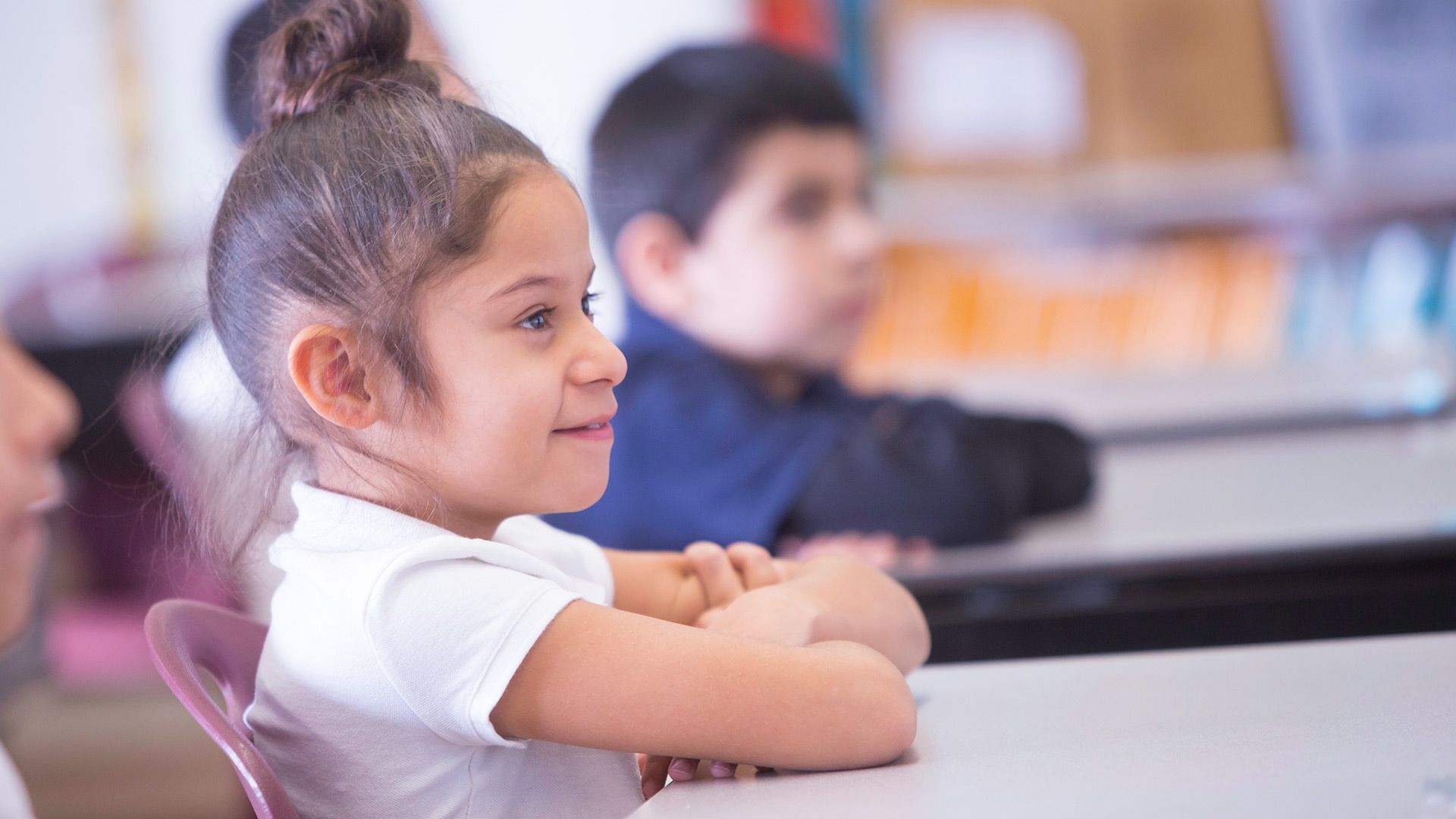 A little girl is sitting at a desk in a classroom with her arms crossed.