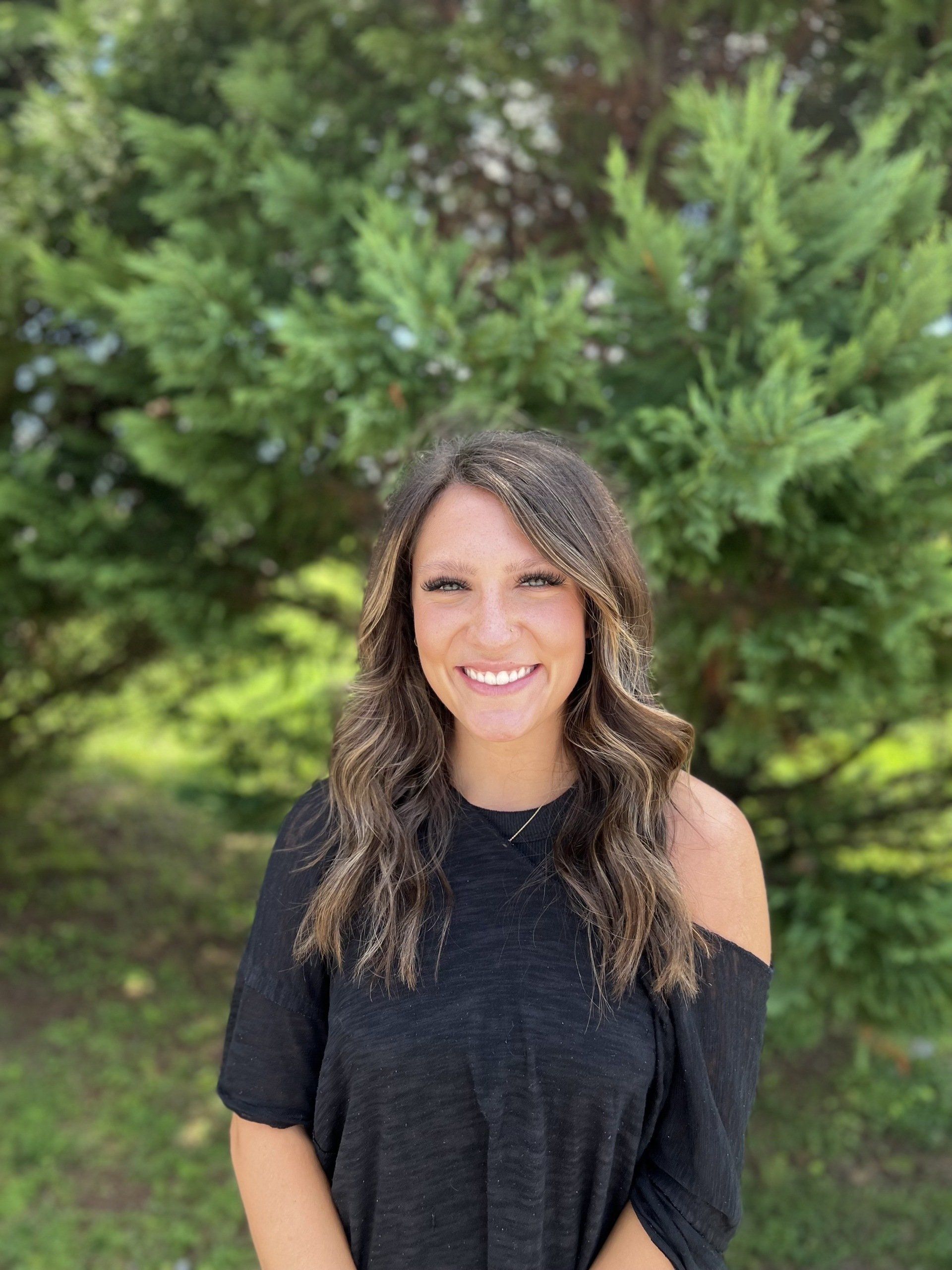 A woman in a black shirt is smiling in front of a tree.