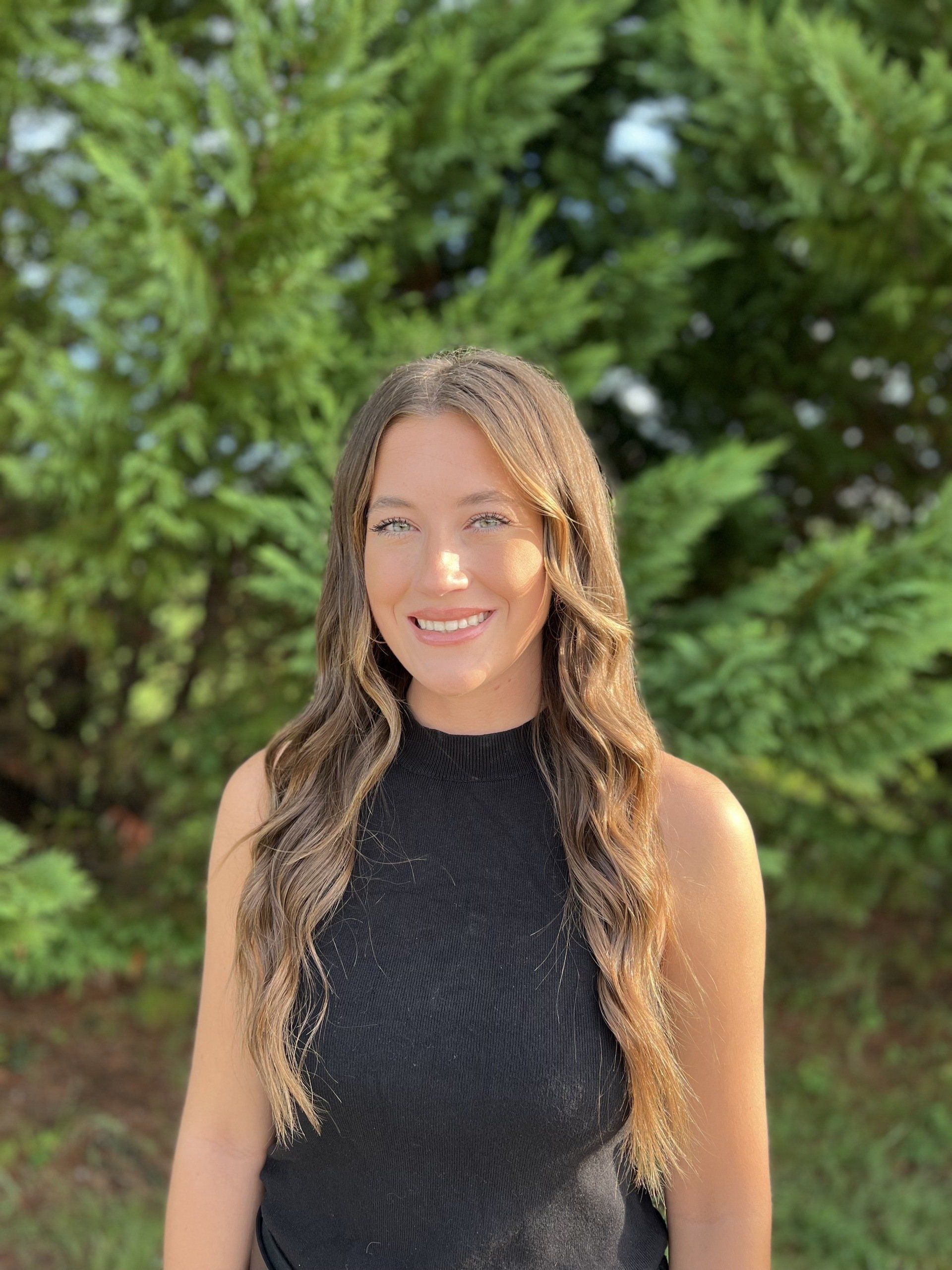 A woman in a black tank top is smiling in front of a tree.