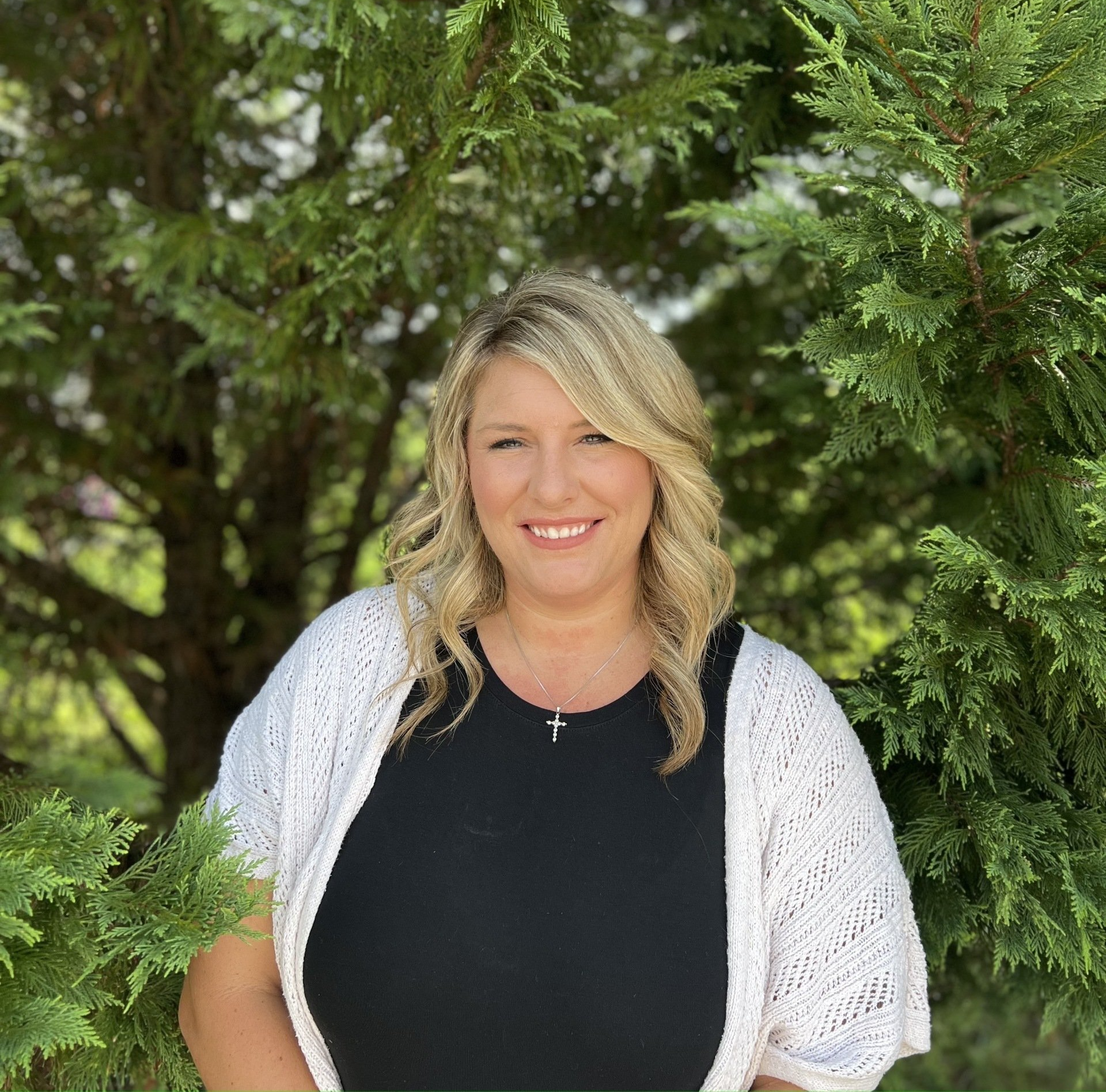 A woman in a black shirt and white cardigan is standing in front of a tree.