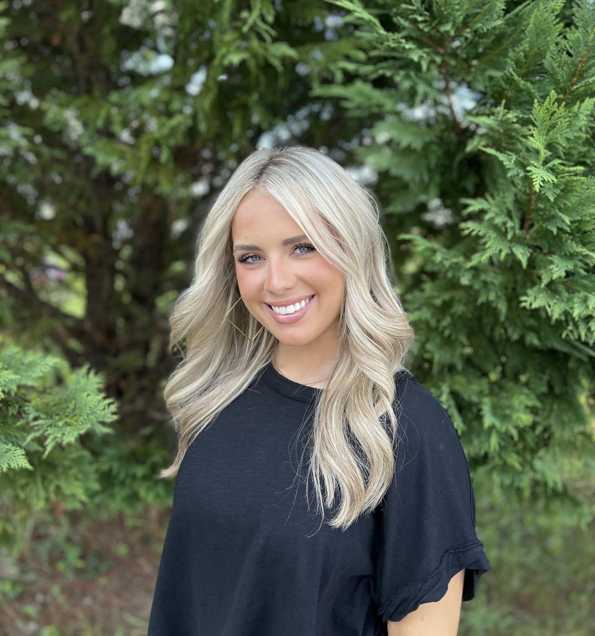 A woman in a black shirt is smiling in front of a tree.