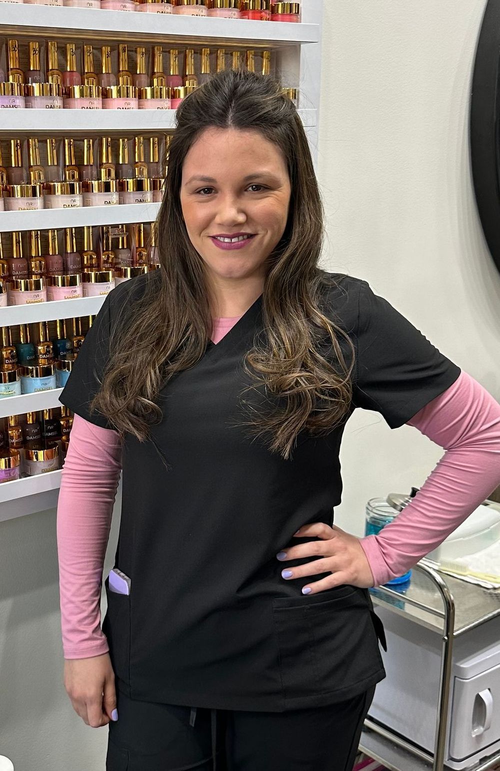 A woman in a black scrub top and pink sleeves is standing in front of a shelf of nail polish.