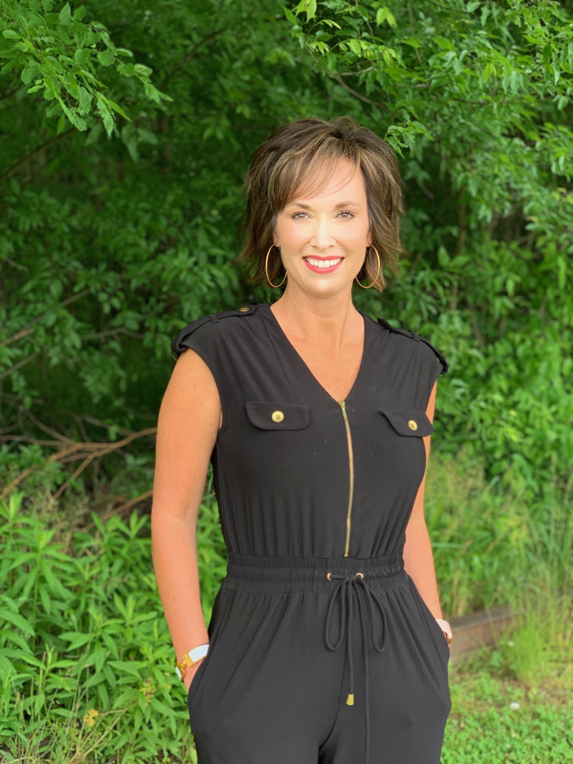 A woman in a black jumpsuit is standing in front of a forest.