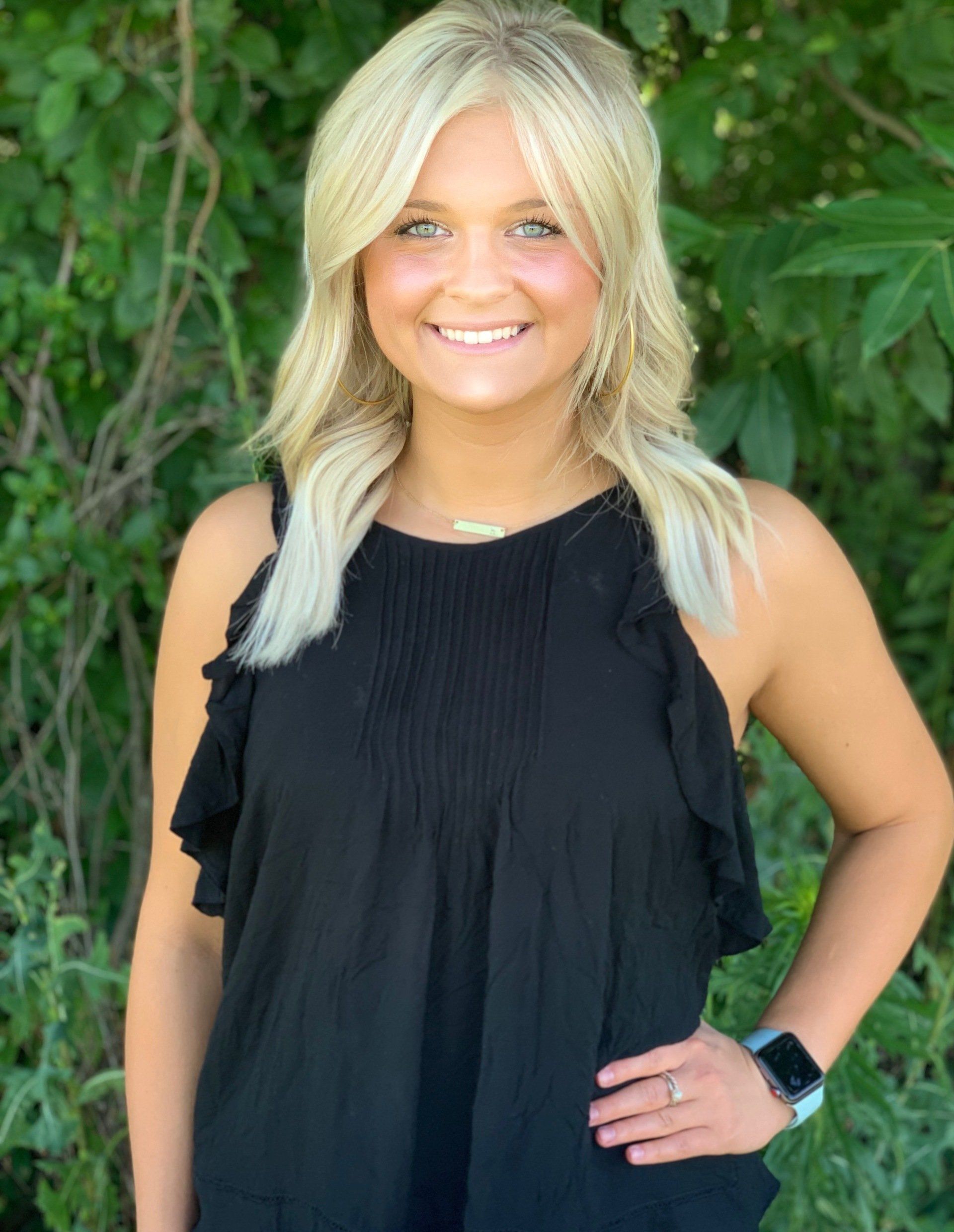 A woman in a black tank top is standing in front of a tree.