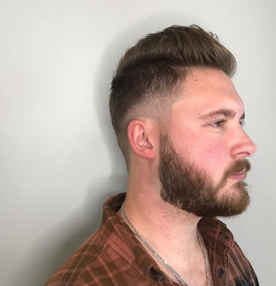 A man with a beard and a plaid shirt is standing in front of a white wall.