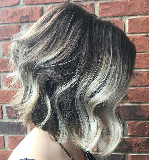 A woman with short wavy hair is standing in front of a brick wall.
