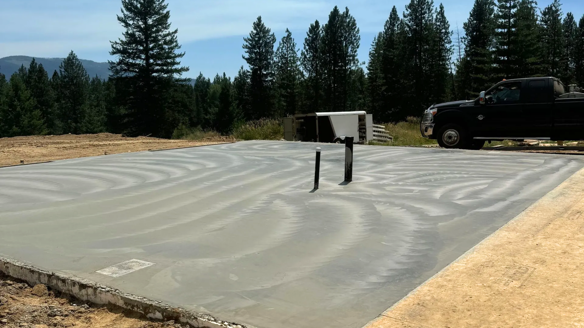 Freshly poured concrete slab foundation with visible plumbing pipes on a rural construction site.