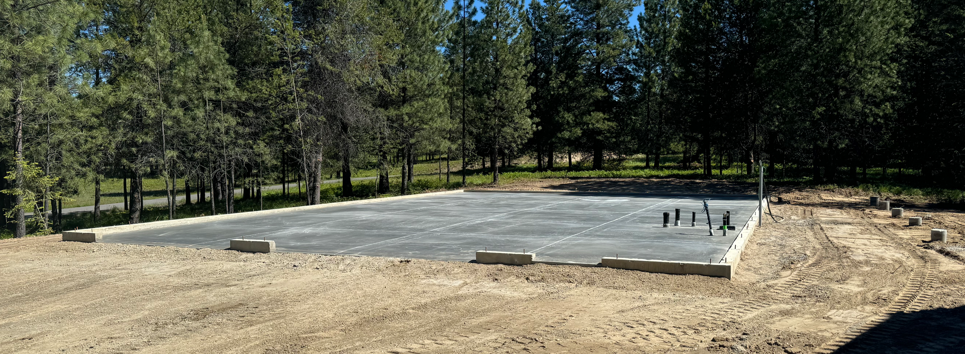 A large concrete slab is sitting in the middle of a dirt field surrounded by trees. Project done by Raise the Bar Construction