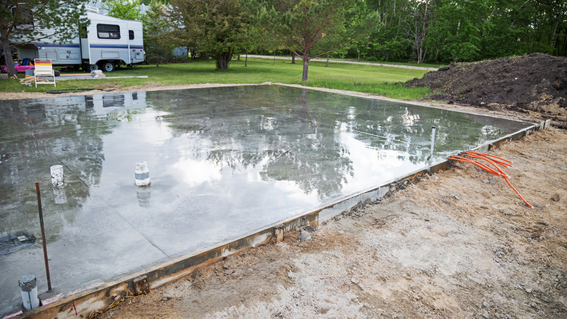 Freshly poured residential concrete slab foundation with plumbing and electrical conduits, set in a grassy yard with surrounding dirt work.