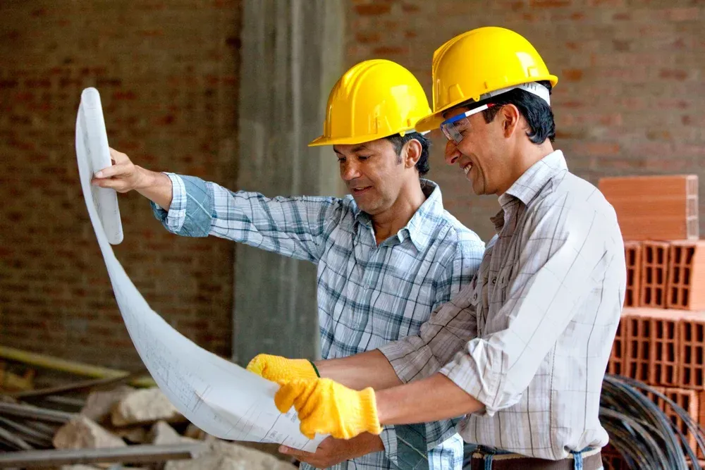 Two construction workers in yellow hard hats examine blueprints at a construction site.