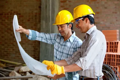 Two construction workers in yellow hard hats examine blueprints at a construction site.