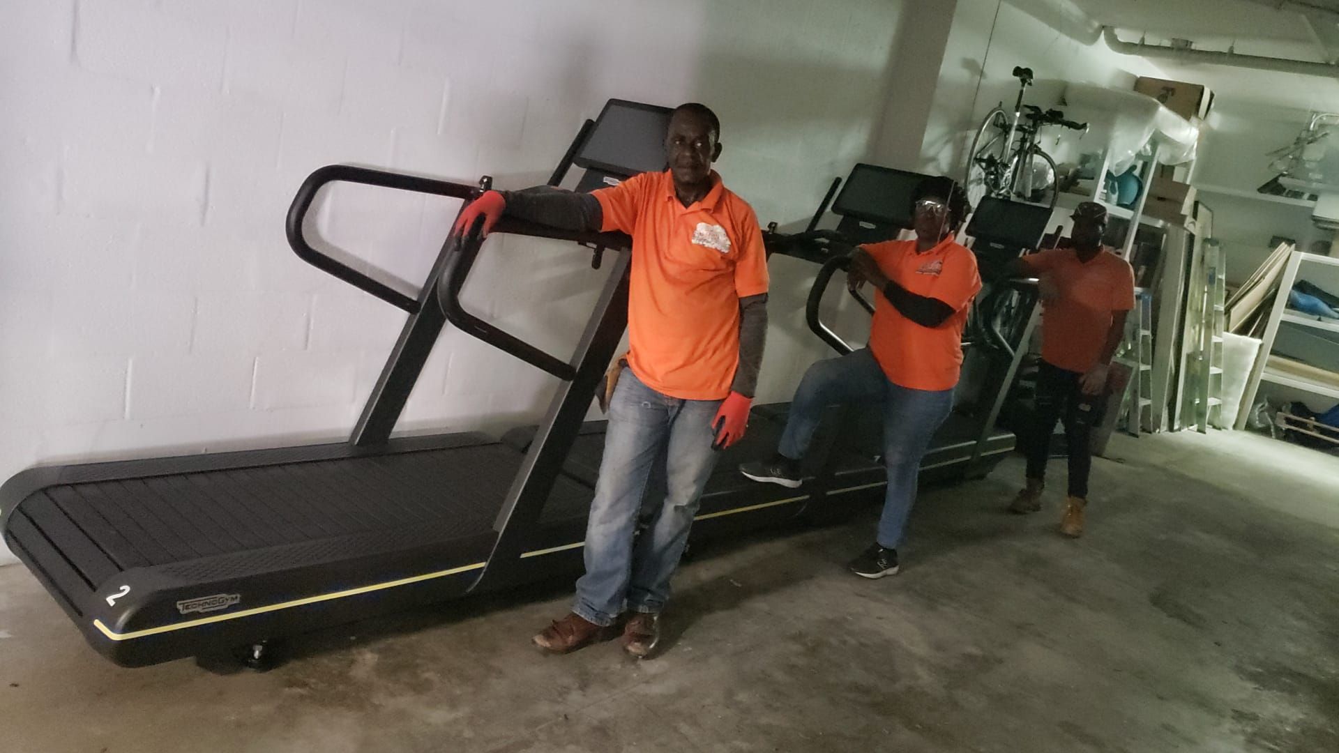 Two men are standing next to a treadmill in a garage.