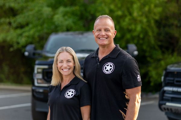 Two people smiling, wearing black shirts with logo, in front of black trucks, outside.