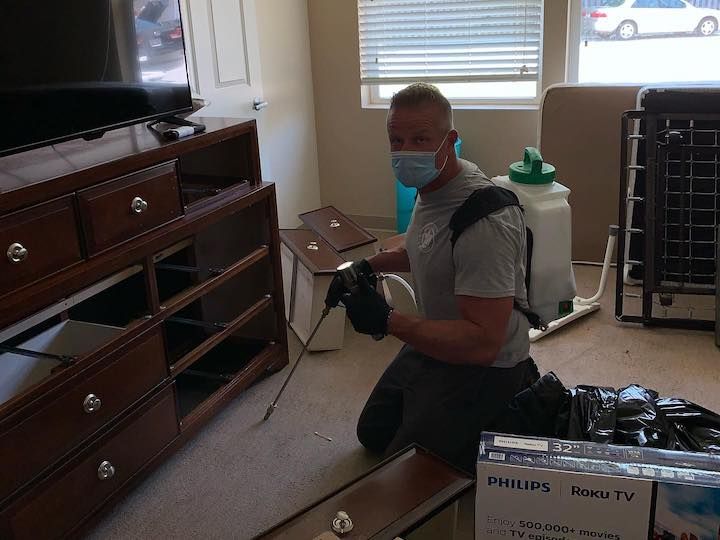 Man wearing mask kneeling, spraying dresser drawers, room interior.