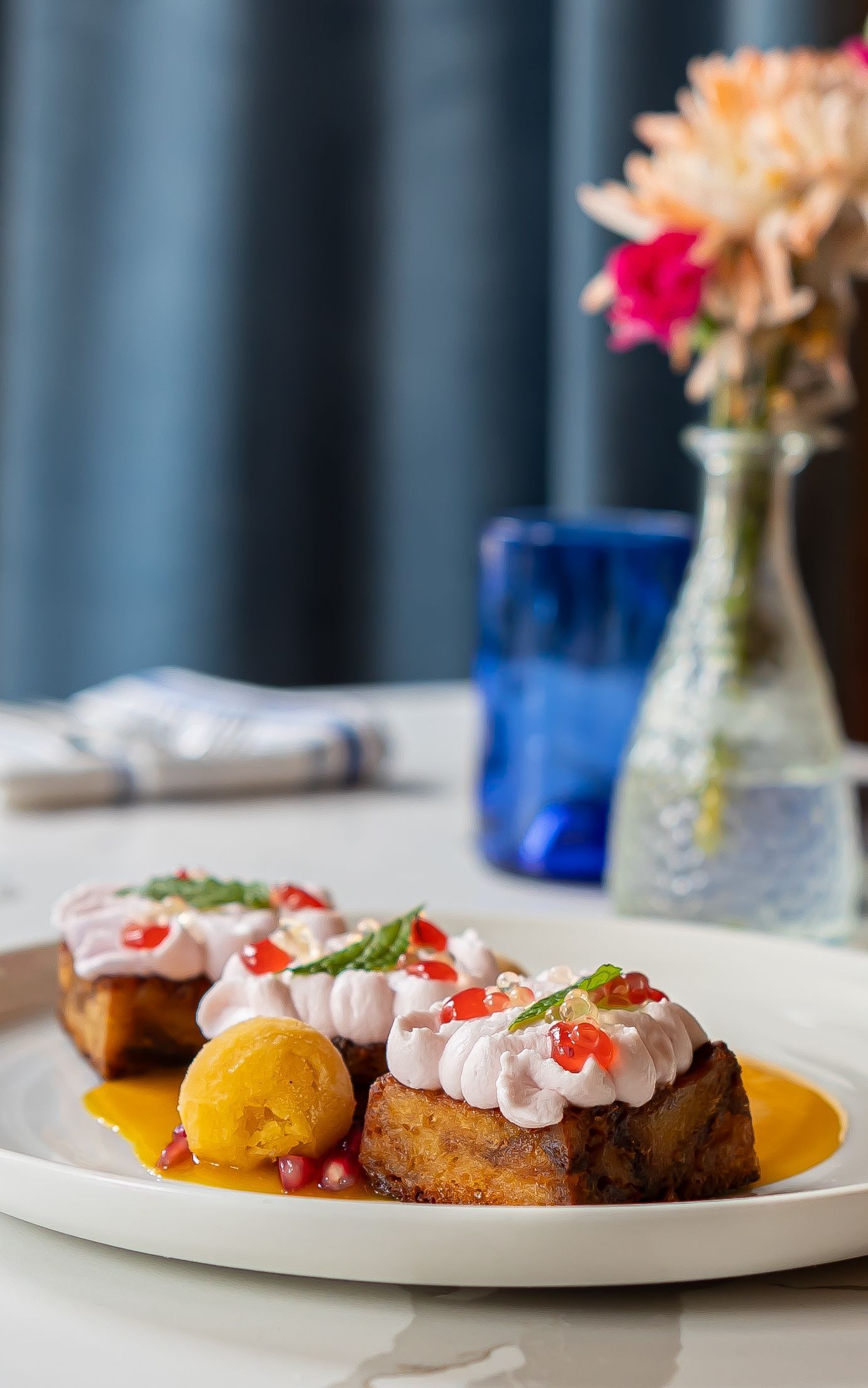 Plate of food: French toast with pink cream and pomegranate, with a side of orange sauce and flower vase in background.