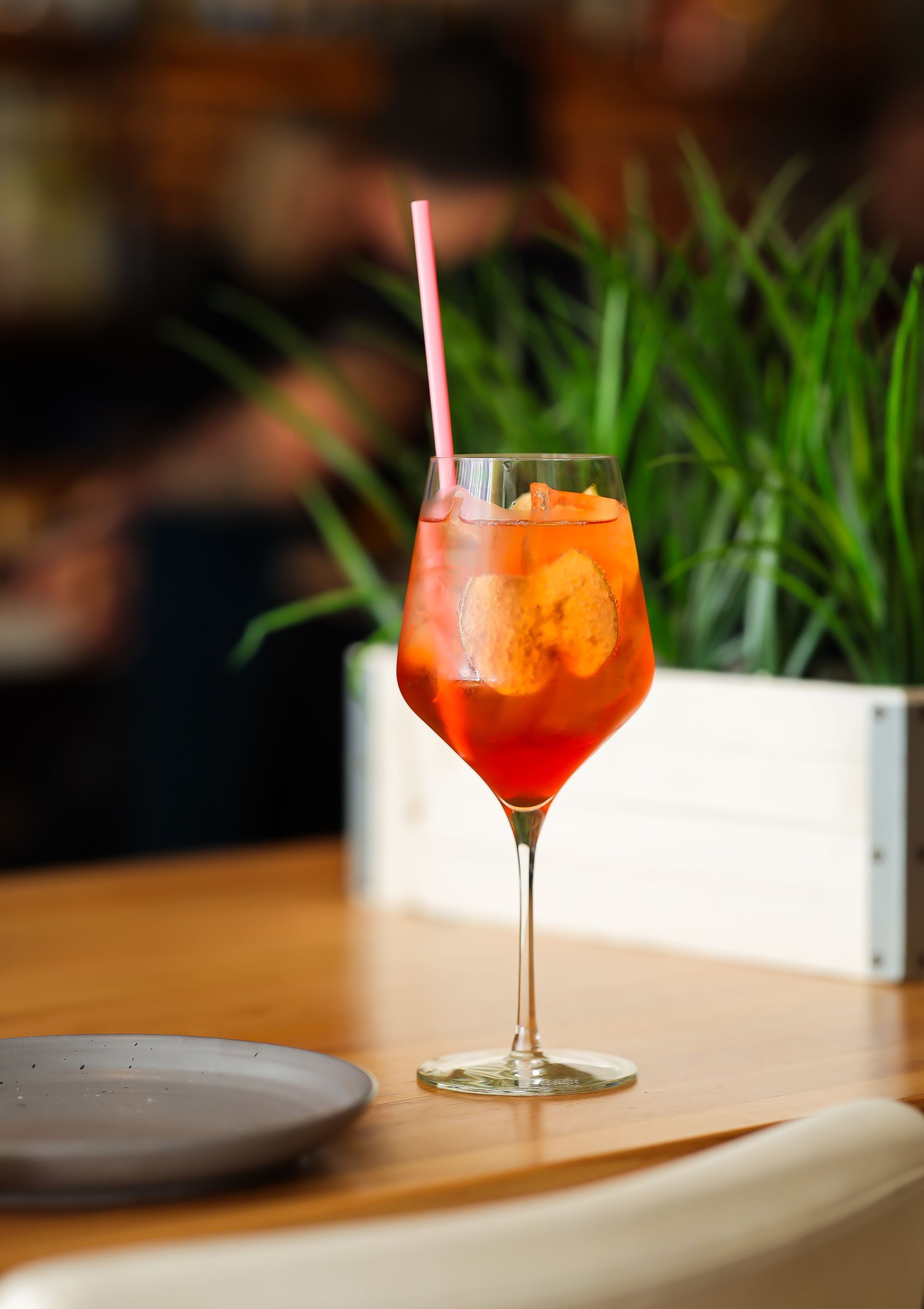 Cocktail in a stemmed glass with orange slices and a pink straw on a wooden table.