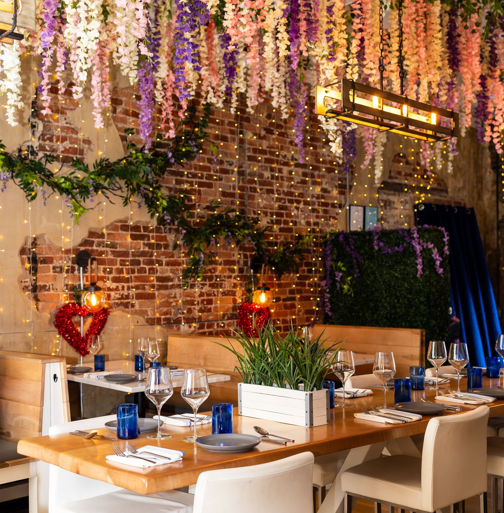 Restaurant interior with a decorated brick wall, tables set for dining, and hanging flowers.