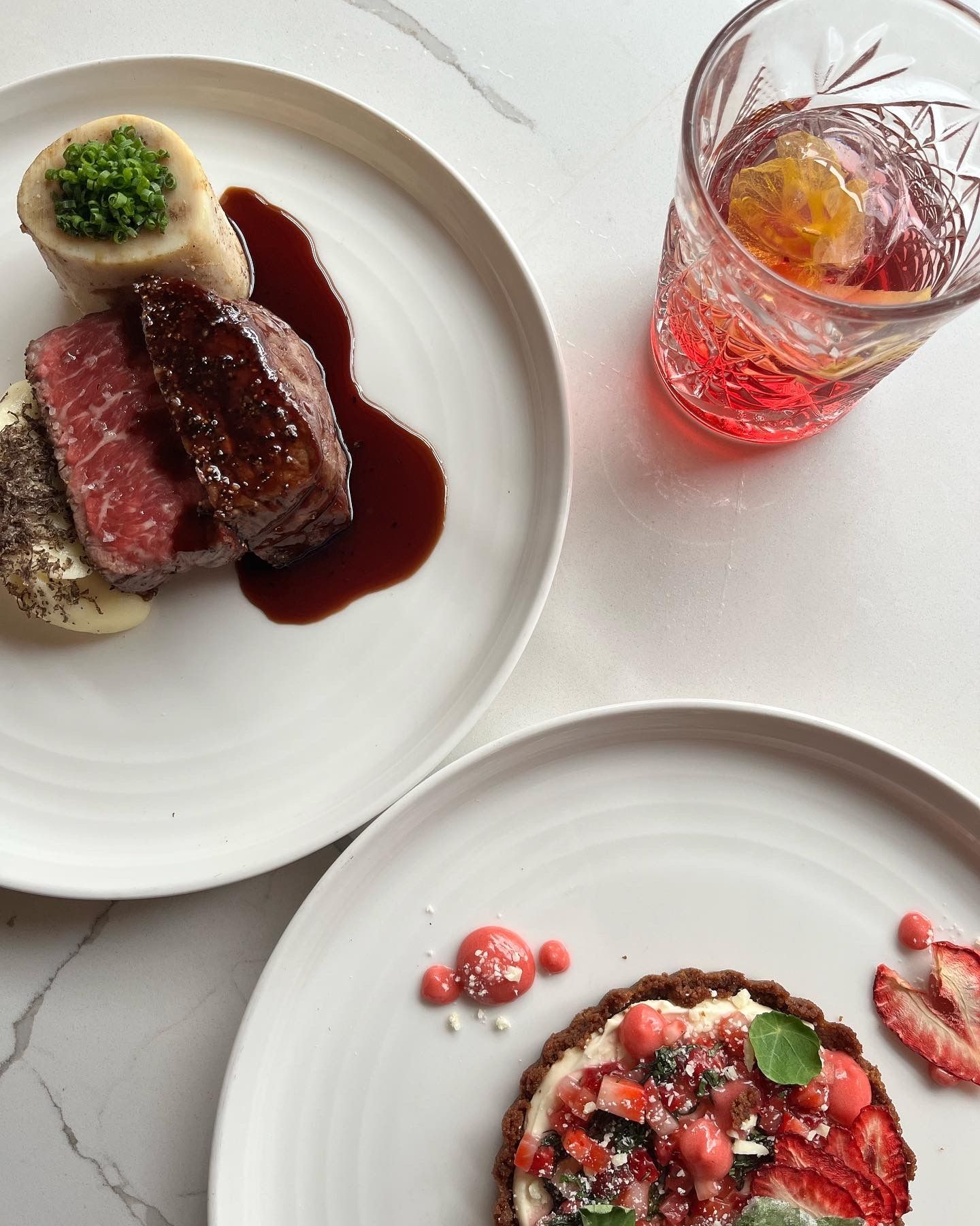 Two plates of food with a cocktail on a white surface. Steak, mashed potato, and tart.