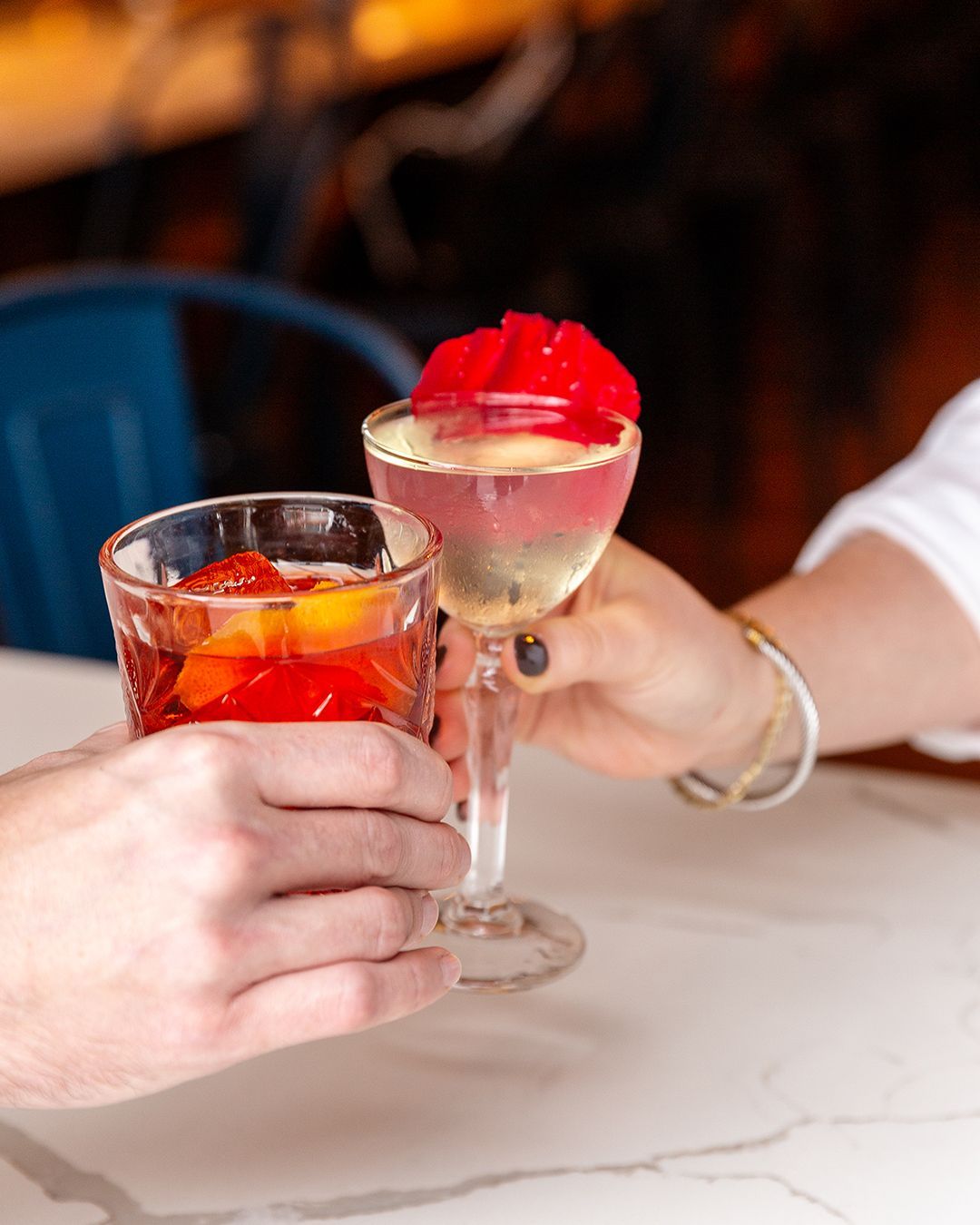 Two people toasting cocktails: a red cocktail in a rocks glass and a pink cocktail with a red garnish in a stemmed glass.