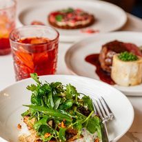 Plated food, including a dish with herbs, a steak with sauce, and a tart. Drinks in glasses.