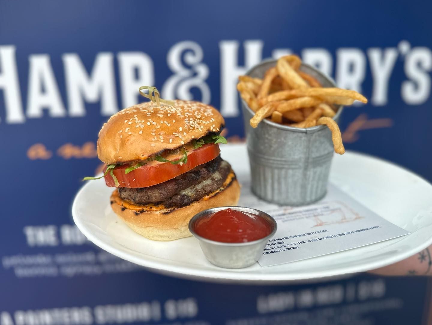 Hamburger with fries and ketchup on a plate in front of a blue sign with restaurant logo.