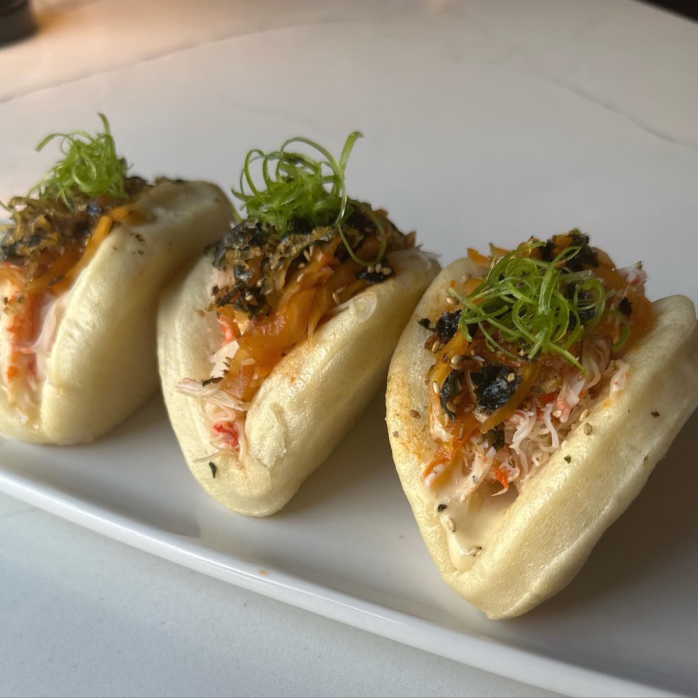 Three bao buns, filled with crab and sauce, topped with green garnish on a white plate.