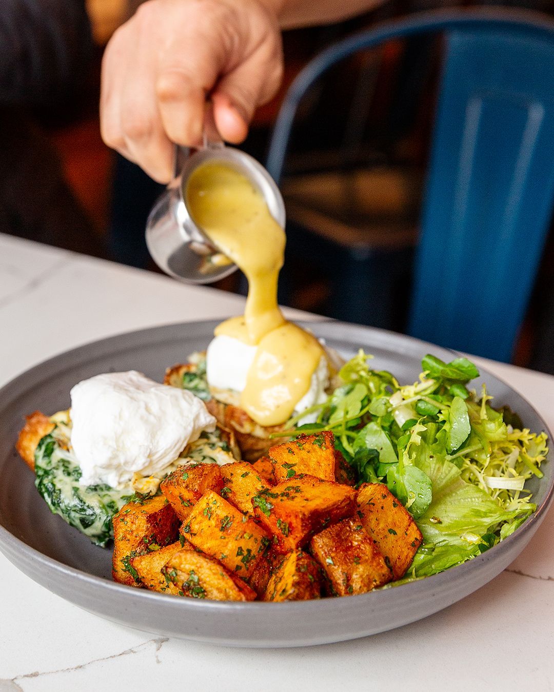 Eggs Benedict with sweet potatoes, salad, and hollandaise sauce being poured.