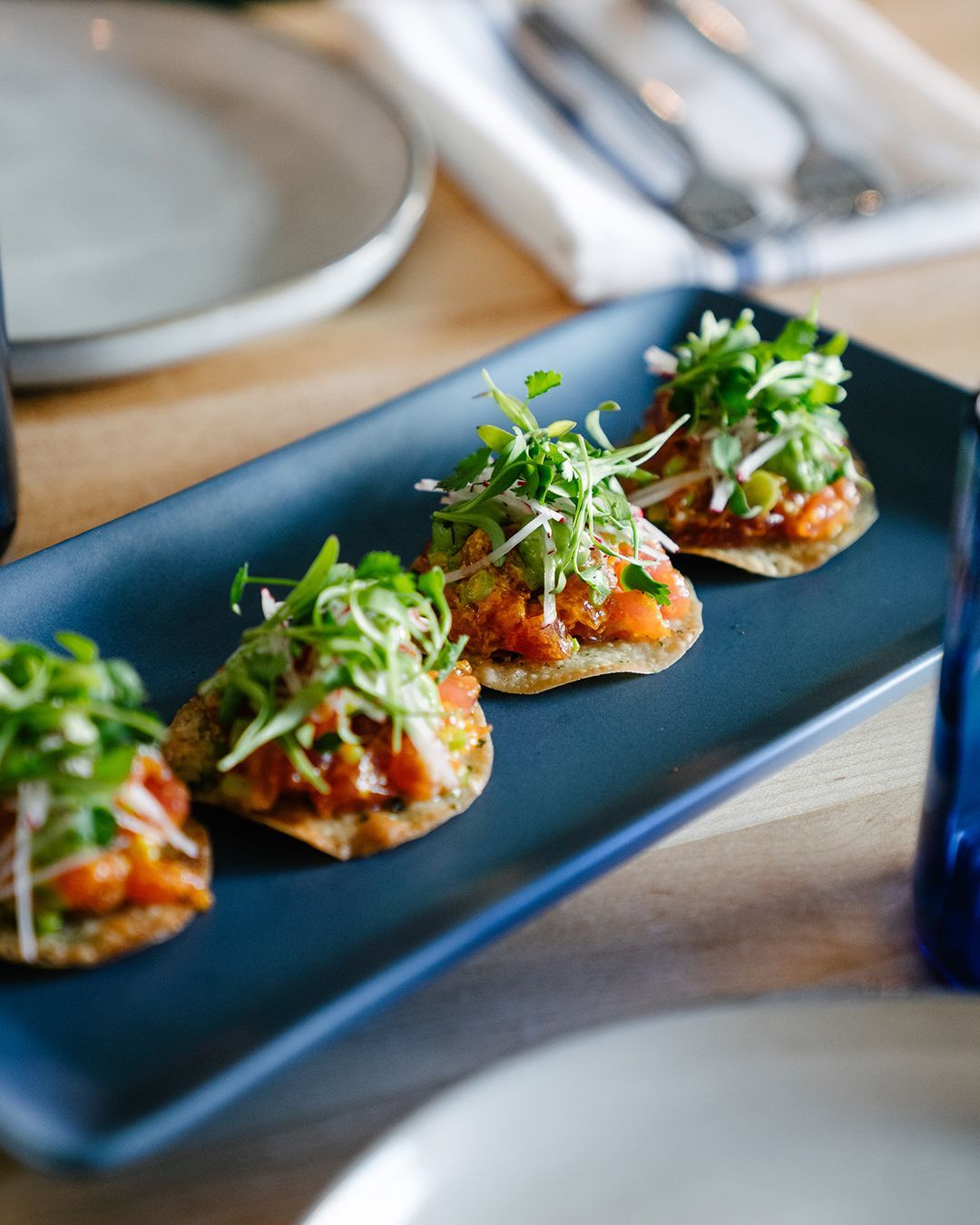 Four appetizers on a blue rectangular plate: fried tortillas topped with salmon, onions, and greens.