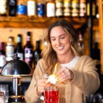 Bartender squeezing orange peel over a red cocktail; lit bar setting.