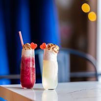 Two colorful cocktails with toppings and straws on a white table. Blue backdrop.