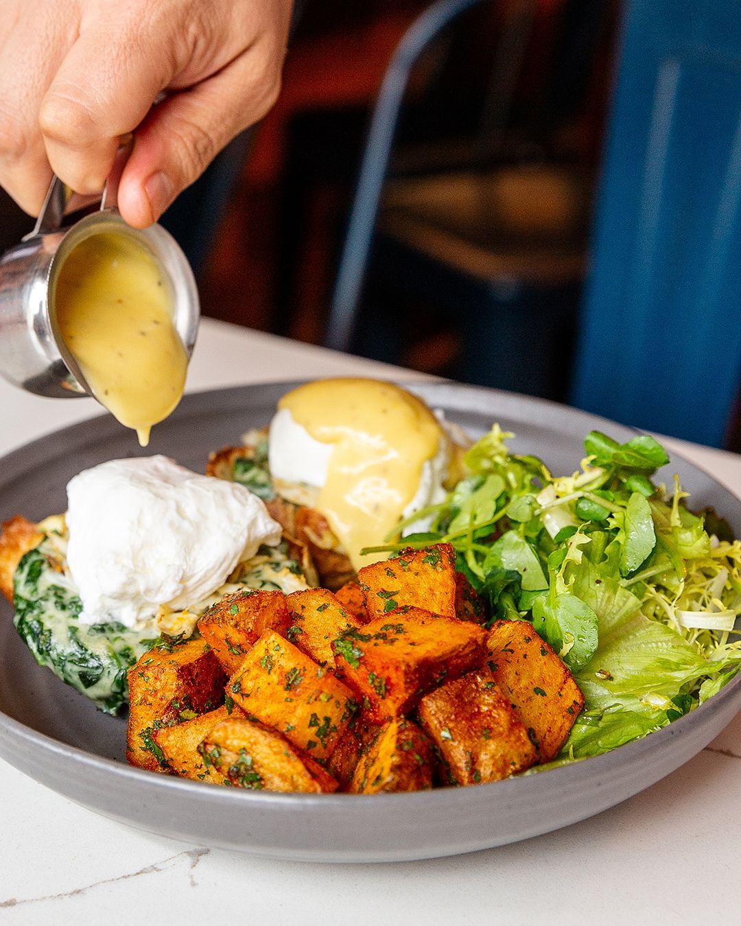 Close-up of a plate with poached eggs, sweet potatoes, greens, and sauce being drizzled.