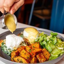 Eggs Benedict with sweet potato hash and greens, being drizzled with sauce.