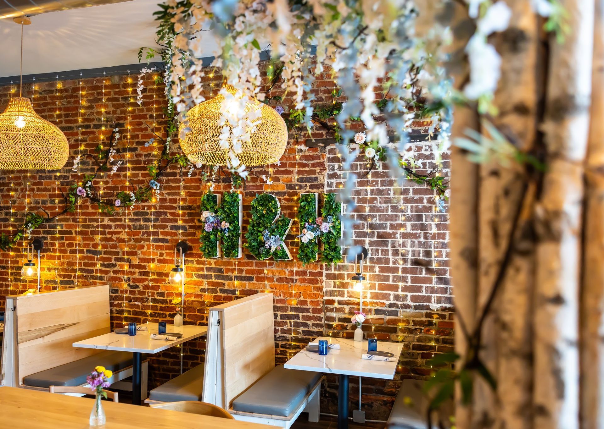 Restaurant interior: exposed brick wall, floral decorations, wicker pendant lights, booths with tables.