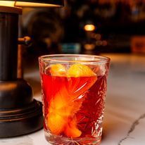 A cocktail in a patterned glass with an orange peel garnish, next to a lamp on a bar.
