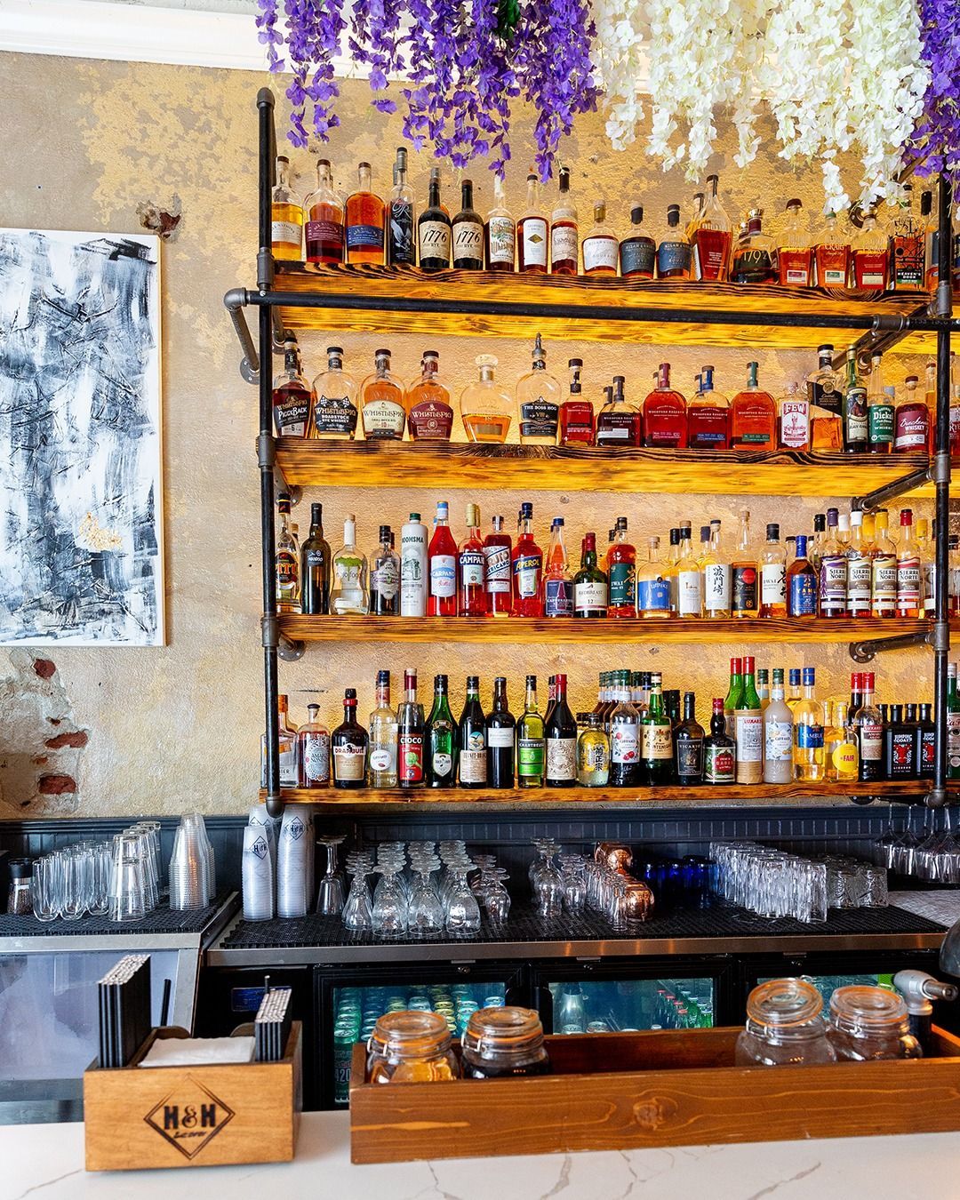 Bar with liquor bottles on shelves, glasses, and purple/white hanging flowers.